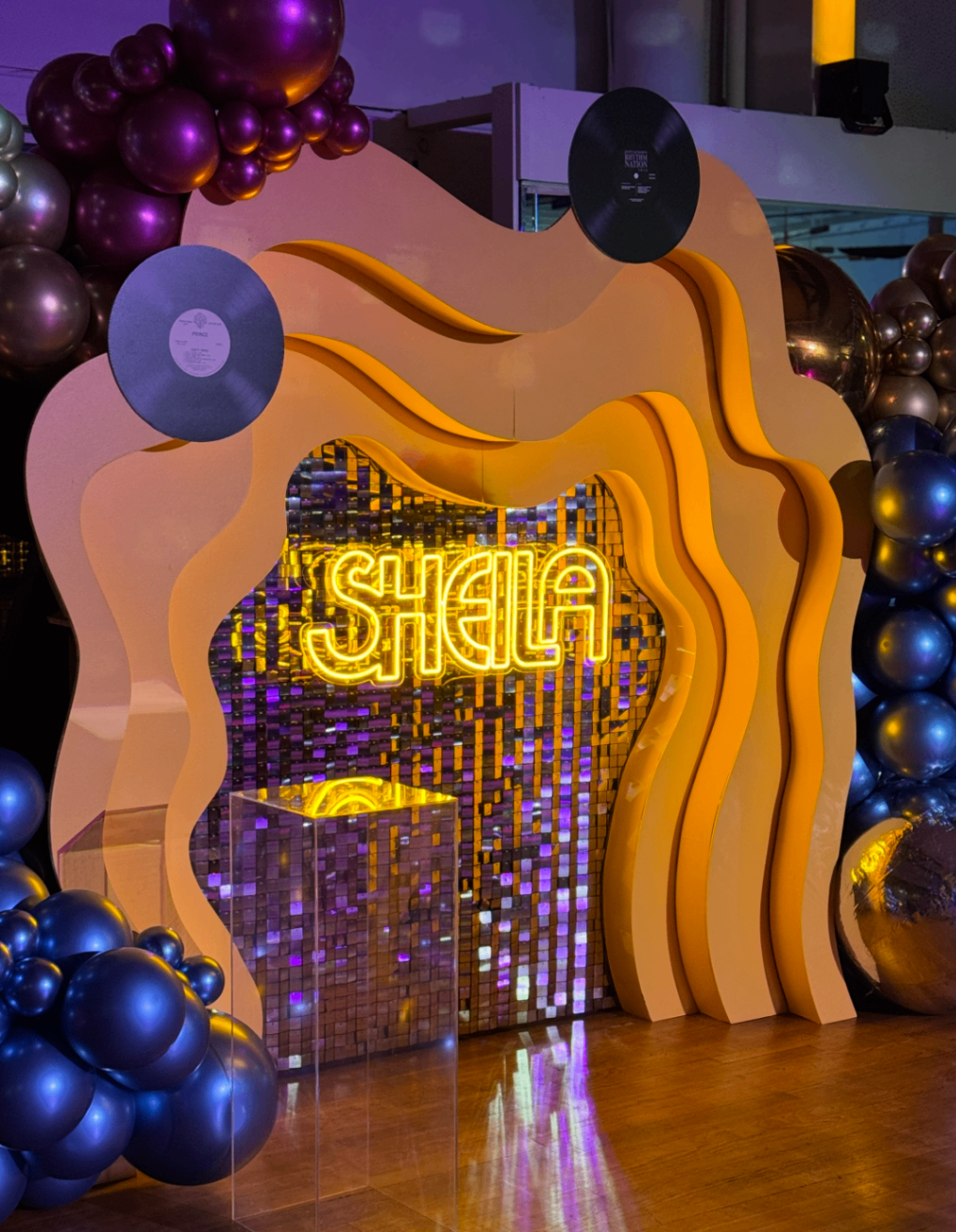 Custom wavy backdrop with neon SHEILA sign, vinyl records, and chrome balloon garlands