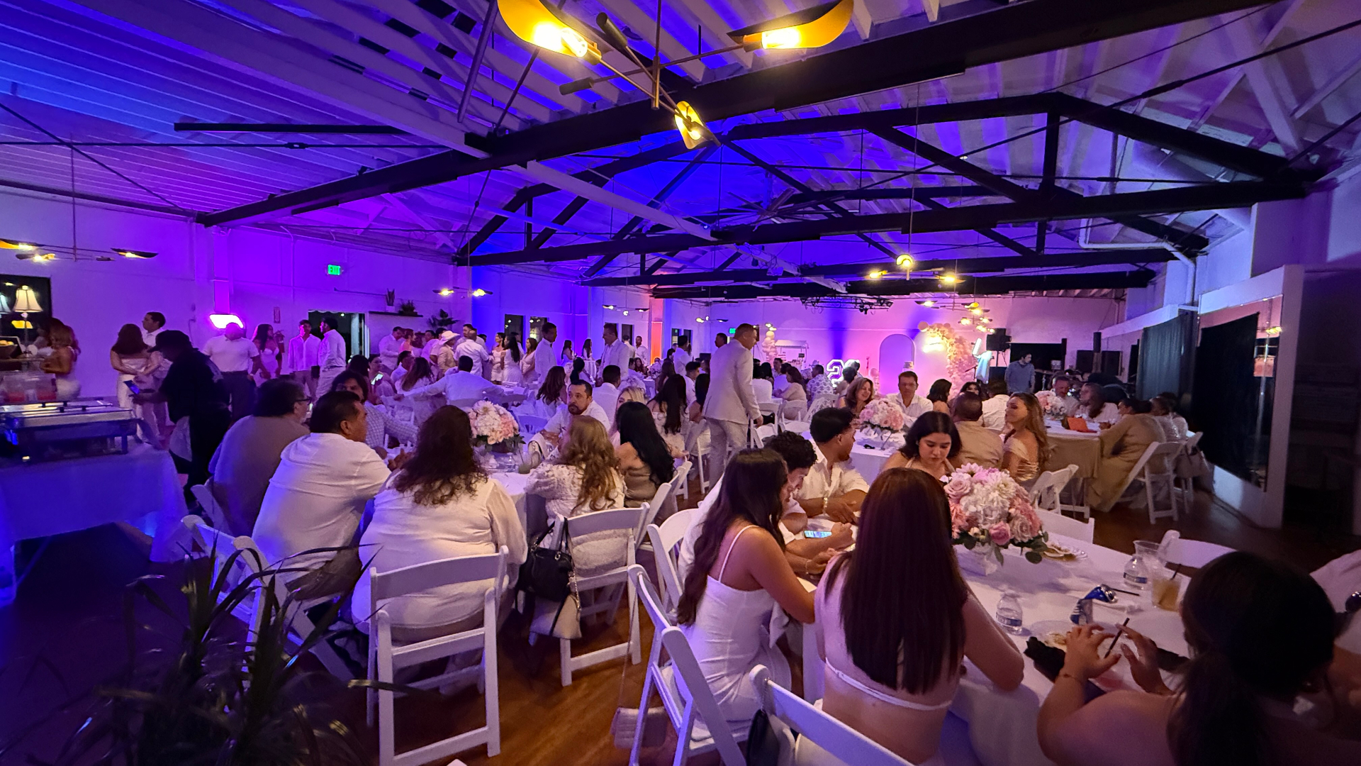 All-white party with guests seated at long banquet tables under purple uplighting and exposed trusses